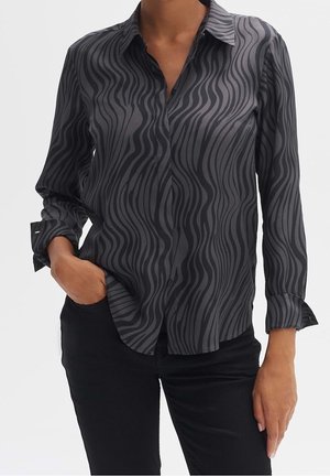 Kobieta ubrana w czarną koszulę na guziki z długim rękawem z abstrakcyjnymi, falistymi szarymi paskami, w połączeniu z czarnymi spodniami.