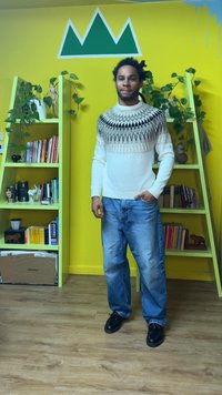 Λευκό πουλόβερ με μαύρα και γκρι γεωμετρικά μοτίβα, συνδυασμένο με ανοιχτό μπλε τζιν και μαύρα παπούτσια. Φωτεινό κίτρινο φόντο με ράφια.