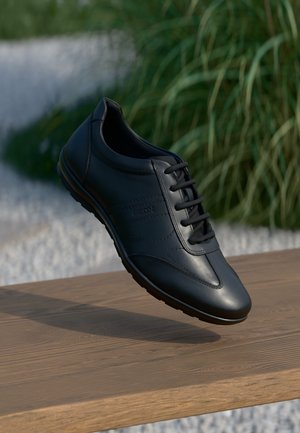 Zwart leren veterschoen voor dagelijks gebruik zwevend boven een houten oppervlak met op de achtergrond vage groene planten en witte stenen.