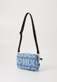 Niebieska torba typu crossbody z denimu z nadrukowanym logo "HUGO". Posiada czarny regulowany pasek, zamek błyskawiczny i srebrne okucia. Prostokątny kształt.