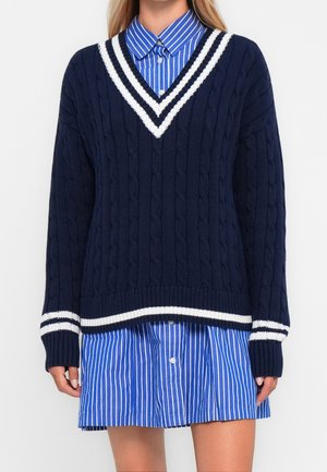 Kobieta ubrana w granatowy sweter w splot warkoczowy z dekoltem w serek i białymi paskami, nosząca pod nim niebiesko-białą sukienkę na guziki w pionowe paski.