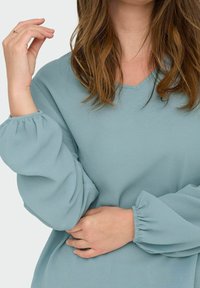 Svijetloplavi V-izrez bluzon s dugim napuhanim rukavima izrađenim od teksturirane tkanine. Kupovljeni manžeti i opušteni kroj su istaknute značajke dizajna.