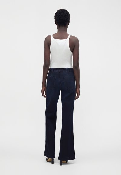 Personne aux cheveux courts et bouclés se tient de dos, portant un haut blanc sans manches, un jean large foncé et des chaussures noires à talons.
