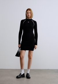 Μαύρο φόρεμα με μακριά μανίκια και λευκές λεπτομέρειες, εφαρμοστό στυλ, με ραβδωτή υφή. Το μοντέλο φοράει μαύρα μοκασίνια και κρατάει μια μικρή μαύρη τσάντα.