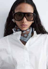Femme aux longs cheveux noirs portant de grandes lunettes de soleil écaille de tortue, des boucles d'oreilles argentées en créole, une chemise blanche à col à volants et une écharpe marron à motifs.