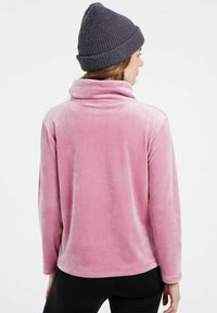 Pullover in velluto rosa con colletto alto e maniche lunghe. Texture liscia con una leggera lucentezza. Indossato con un berretto lavorato a maglia scuro.
