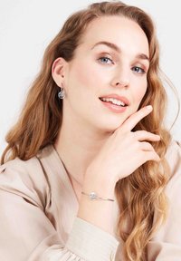 Zilveren hoepel oorbellen met een circulair ontwerp en ingewikkelde details. Eenvoudige zilveren armband met een gecentreerde heldere edelsteen. Zachte beige blouse.