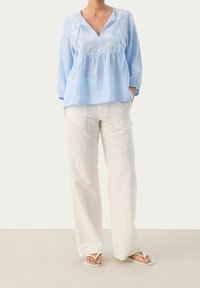 Blusa in lino azzurro chiaro con scollo a V, maniche lunghe e dettagli ricamati bianchi, abbinata a pantaloni larghi crema e infradito.