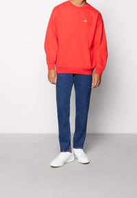 Roter Pullover mit Rundhalsausschnitt und langen Ärmeln, kombiniert mit blauen Jeans und weißen Turnschuhen, mit einem Logo auf der Brust.