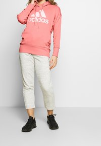 Sweat à capuche rose avec grand logo adidas blanc, associé à un pantalon de jogging court gris clair à motifs et des chaussures de sport noires. Style décontracté et confortable.