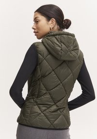 Gilet matelassé vert olive avec capuche, présentant un motif en losanges et une finition légère et brillante. Porté par-dessus un haut noir à manches longues.