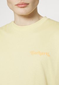 Yellow short-sleeve t-shirt made from cotton featuring a subtle orange "Carhartt" logo on the chest. Smooth texture, classic fit.