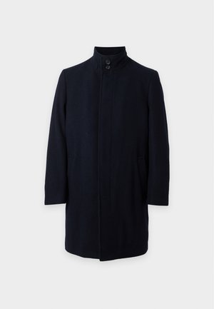 Homme portant un chapeau bob noir, un manteau noir, une chemise blanche, un pantalon marron à carreaux et des mocassins noir et blanc, debout les mains dans les poches.