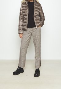 Kurtka puchowa w metalicznie brązowym kolorze z kapturem wykończonym futrem, zestawiona z beżowymi spodniami w kratę i czarnymi masywnymi botkami na kostkę. Pod spodem strukturalny sweter dzianinowy.