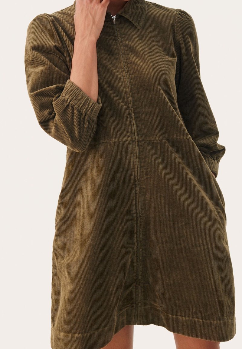 Femme portant une robe en velours côtelé vert olive avec une fermeture éclair sur le devant et des manches trois-quarts, debout devant un fond uni.