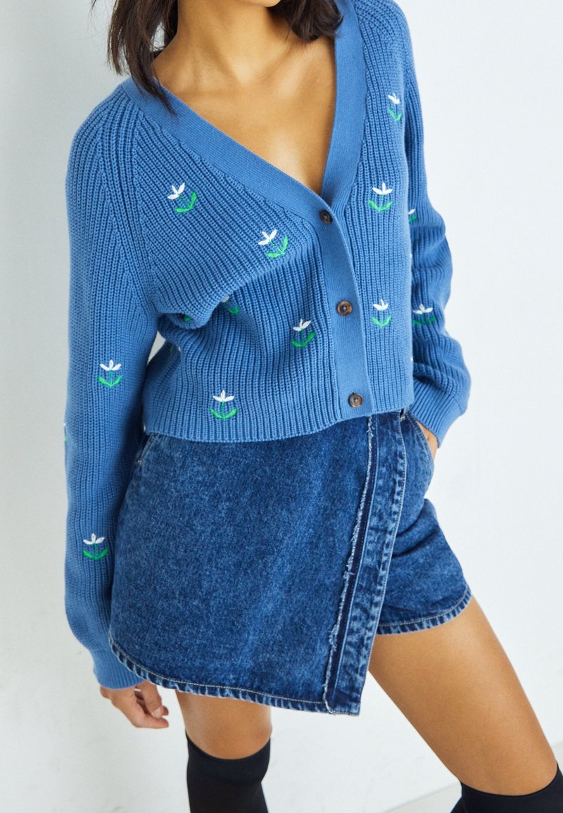 Cardigan en tricot bleu avec un col en V, orné de broderies florales blanches et vertes. Associé à une mini-jupe en denim à fente.