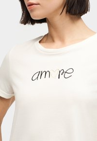 Γυναίκα που φορεί λευκό T-shirt με τη λέξη «amore» σε μαύρα καλλιγραφικά γράμματα, με μια χρυσή καρδιά να αντικαθιστά το γράμμα «o».
