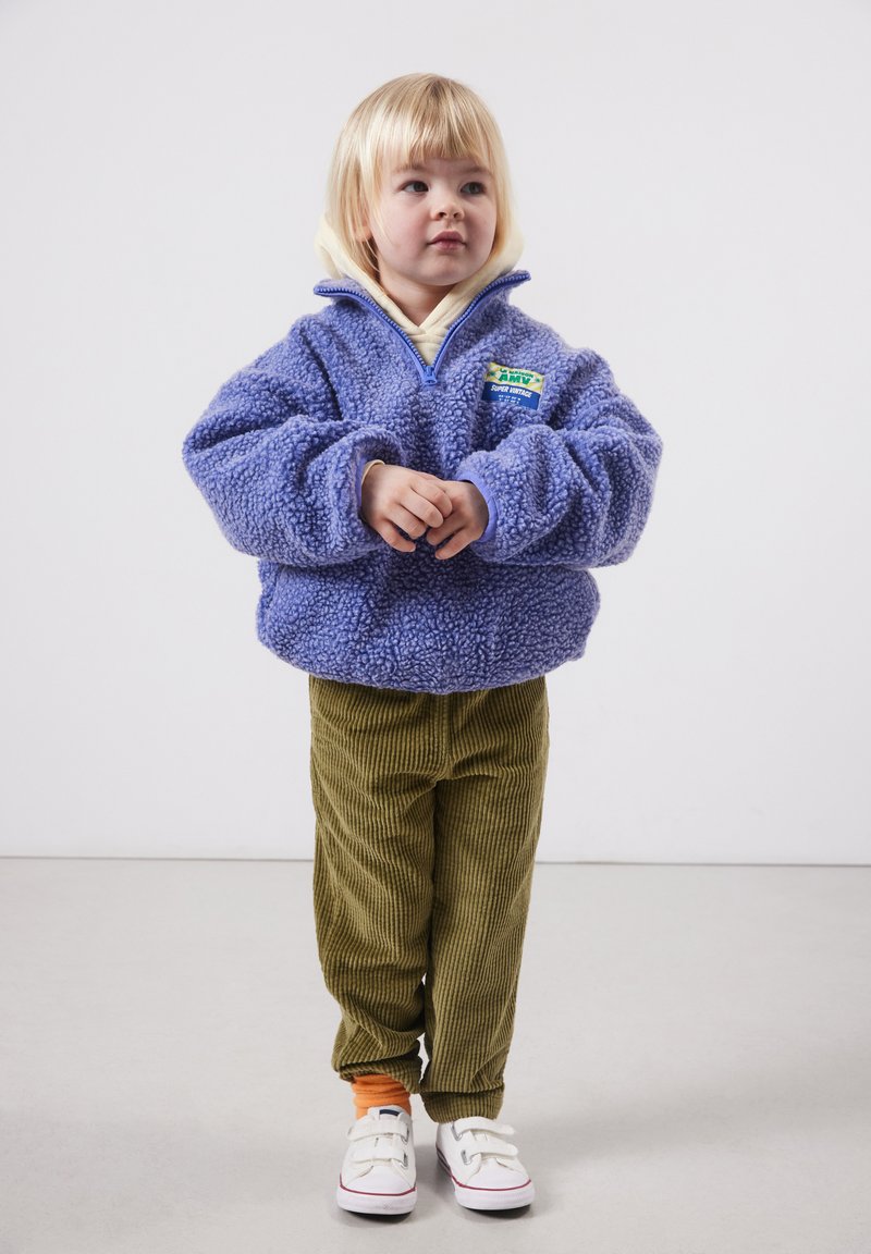 Tout-petit aux cheveux blonds portant une veste duveteuse violette, un pantalon en velours côtelé vert olive, des chaussettes orange et des baskets blanches, debout sur un sol uni.