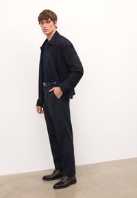 Giacca in suede nera, camicia blu navy, pantaloni scuri su misura e scarpe in pelle marrone. Il modello si trova in un ambiente dai toni neutri. Design minimalista.