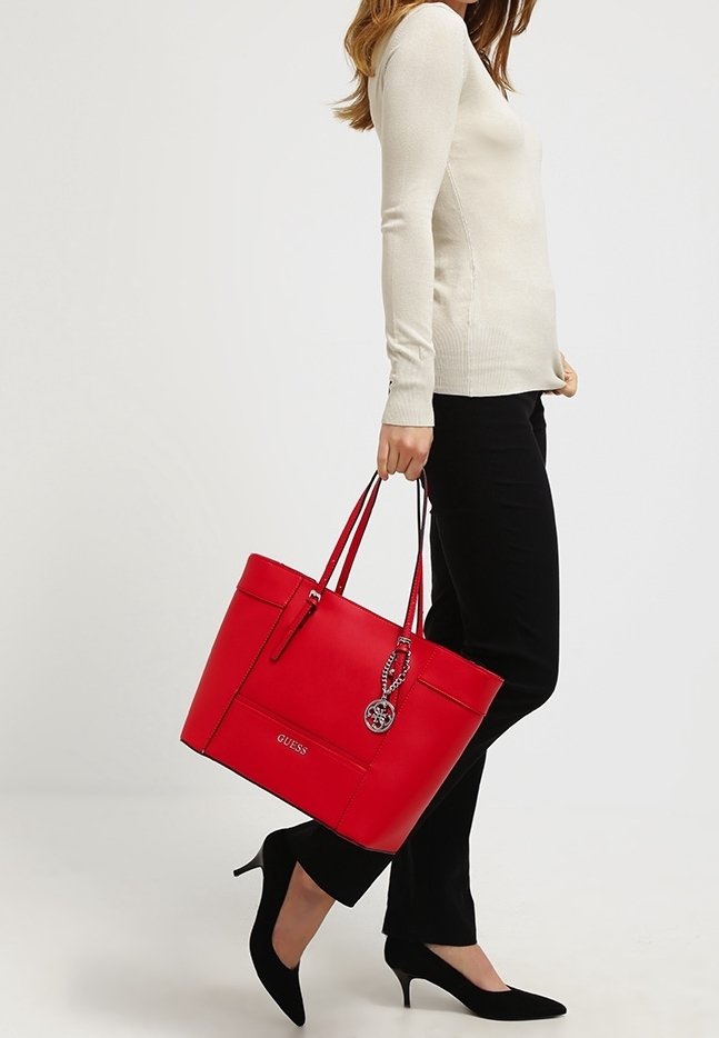 Femme tenant un grand sac fourre-tout rouge Guess, portant un haut blanc à manches longues, un pantalon noir et des chaussures à talons noirs, marchant sur un fond blanc.