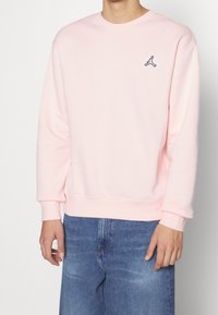 Sudadera rosa con cuello redondo y puños de canalé. Presenta un pequeño logo azul marino en el lado izquierdo del pecho. Combinada con jeans de mezclilla azules.