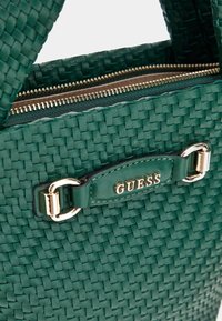 Zöld, szőtt kézitáska arany cipzárral és bőr címkével, aminek a "GUESS" logója van rajta. Kiemelkedik a textúrált felületével és a fém kiegészítőivel.