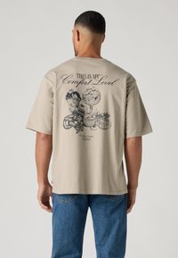 Smėlio spalvos medvilninė marškinėliai laisvo kirpimo. Ant nugaros yra desertų grafikos atspaudas ir užrašas „THIS IS MY Comfort Level“.