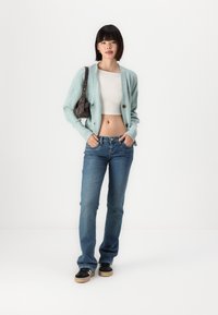 Jasnoniebieski sweter dziergany z guzikami, biały top crop, niebieskie dżinsy i czarne buty z brązowymi podeszwami. Modelka trzyma małą brązową torebkę.