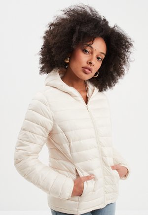 Veste matelassée beige clair avec un design quilté, capuche et fermeture éclair. Comprend des poches latérales et une texture lisse et brillante.