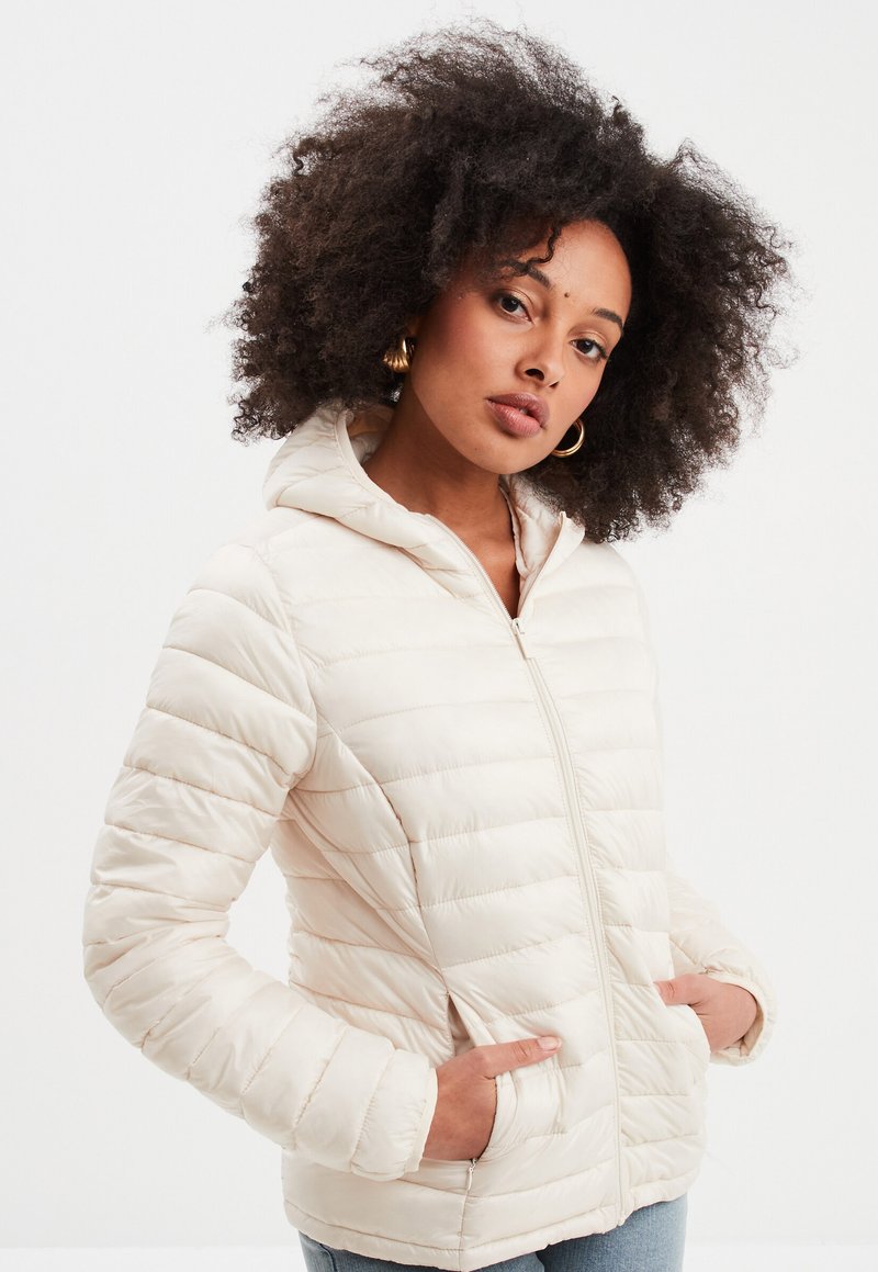Giacca puffer leggera color beige chiaro con design trapuntato, cappuccio e chiusura a zip. Presenta tasche laterali e una texture liscia e lucida.