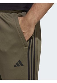 Olijfgroene joggerbroeken met een gladde textuur, voorzien van zwarte zijstrepen en een Adidas-logo nabij de zak voor branding.
