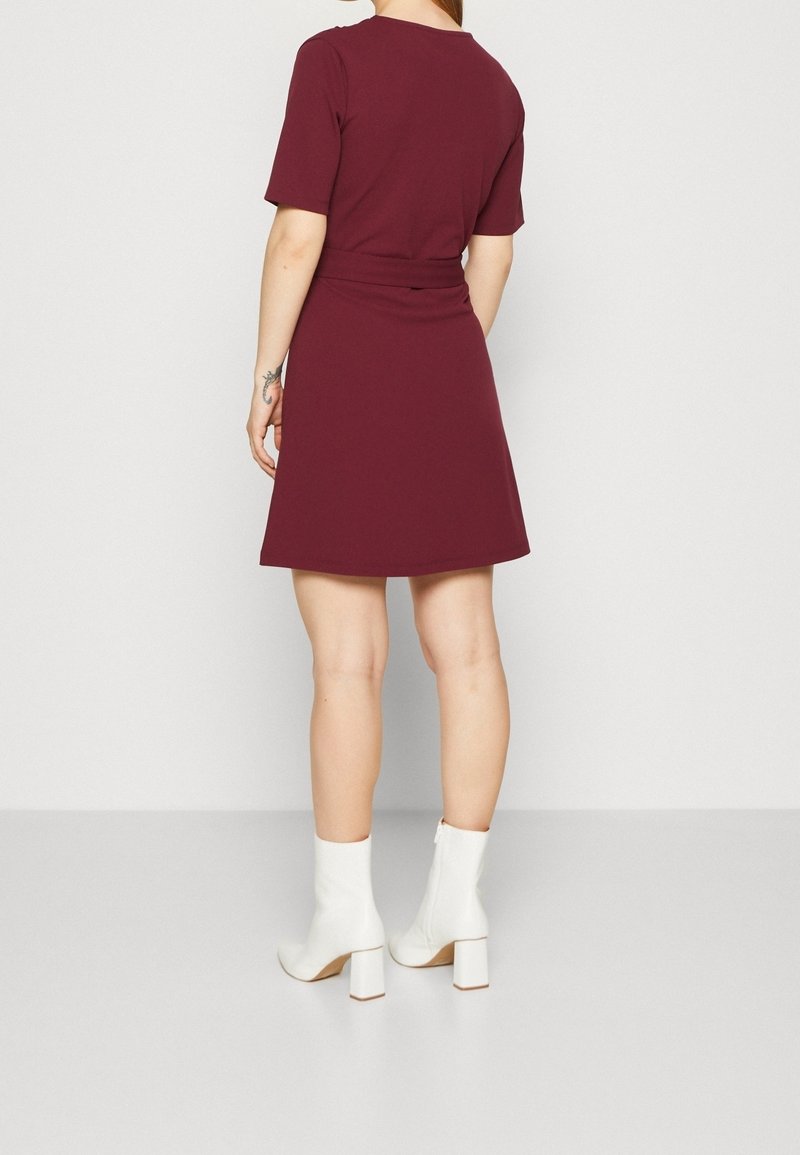 Vestido corto de manga corta en color burdeos con cintura ajustada, falda acampanada y textura suave. Llevado con botas de tobillo blancas de tacón cuadrado.