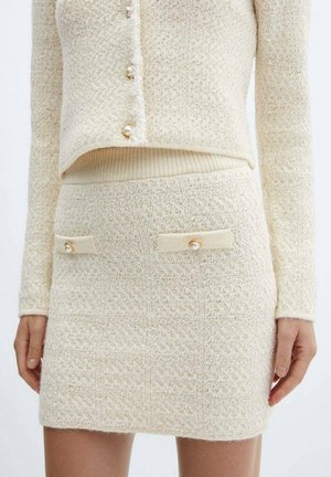 Jupe tricotée texturée crème avec deux poches devant, chacune fermée par un bouton en perle, portée avec un cardigan court assorti.