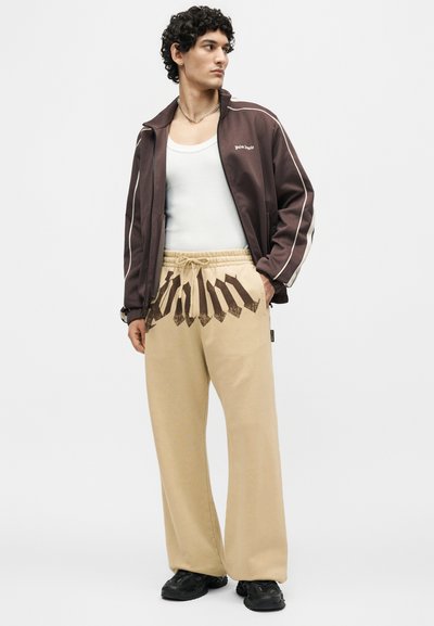 Νέος άνδρας με καφέ ζακέτα με φερμουάρ, άσπρο αμάνικο, μπεζ παντελόνι με μεγάλο μαύρο κείμενο και μαύρα αθλητικά παπούτσια, όρθιος με ένα χέρι στην τσέπη.