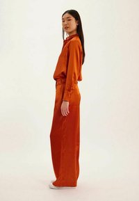 Tenue en satin de couleur rouille, composée d'une chemise ample à manches longues et de pantalons à jambes larges, avec une texture lisse. Des chaussures blanches viennent compléter le look.