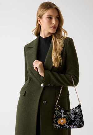 Grüner Wollmischmantel mit Kerbkragen und Knopfdetails. Hält eine schwarze Samttasche mit Blumenstickerei und Kettenriemen.