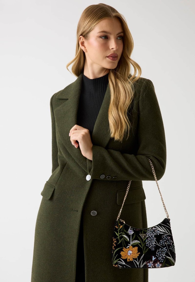 Zielony płaszcz z mieszanki wełny z klapami z wycięciami i dodatkami z guzików. Trzyma czarną aksamitną torbę z kwiatową haftowaną aplikacją i łańcuszkowym paskiem.