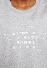 T-shirt gris Adidas avec logo et texte en russe, japonais, chinois et anglais indiquant « "Three Stripe Life" » sur la poitrine.
