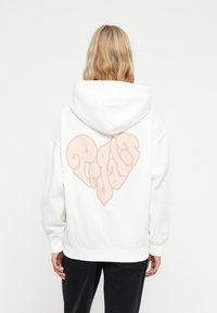 Hvit hettegenser med et stort, mykt rosa hjerte-design og stilig tekst på baksiden. Laget av et tykt, strukturert stoff med hette.
