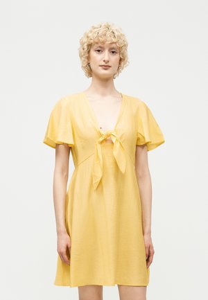 Young person with short curly blonde hair wearing a short-sleeve yellow dress tied in a front knot against a plain background.