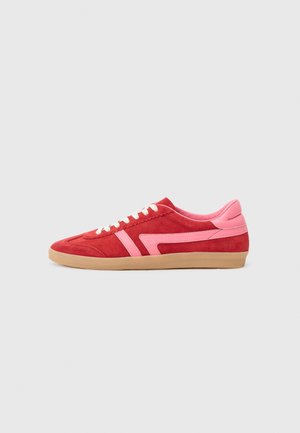 Zapatilla de ante roja con detalles en cuero rosa, cordones blancos y una suela de caucho marrón claro, vista de lado sobre un fondo liso.