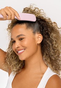 Kvinne med krøllete hår som smiler mens hun børster toppen av hodet med en bredtannet rosa kam mot en ensfarget bakgrunn.