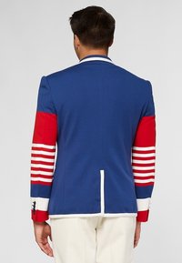 Homme de dos portant un blazer bleu avec des manches rayées rouges et blanches et un pantalon blanc sur un fond uni.