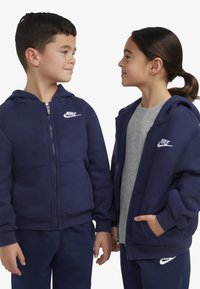 Des sweat-shirts zippés Nike en bleu marine, l'un avec un t-shirt gris en dessous. Les caractéristiques incluent des accents de logo et des poignets côtelés. Matière douce.