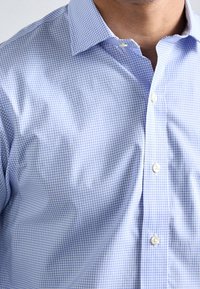 Camicia a quadri azzurro chiaro con colletto button-down. Il tessuto ha una texture liscia e presenta bottoni bianchi lungo la parte anteriore.