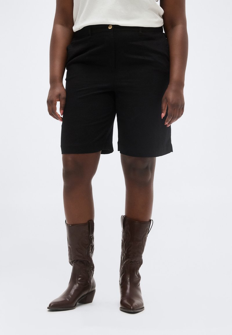 Άτομο που φοράει μαύρα σορτς μέχρι το γόνατο, λευκό μπλουζάκι, και καφέ σκουρά δερμάτινα καουμπόικα μποτάκια μέχρι τη μέση της γάμπας, στέκεται μπροστά σε απλό φόντο.