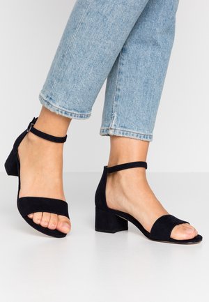 Pieds portant des sandales en daim noir à talon bloc ouvert à l'orteil avec brides à la cheville, associées à un jean court en denim bleu clair.