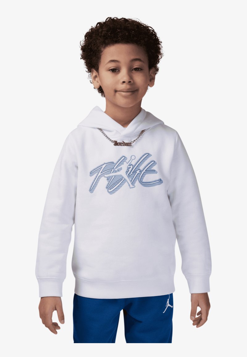 Niño con cabello rizado que lleva una sudadera blanca con el texto "Flight" en azul y pantalones azules con un logo Jumpman en blanco, de pie frente a un fondo blanco.
