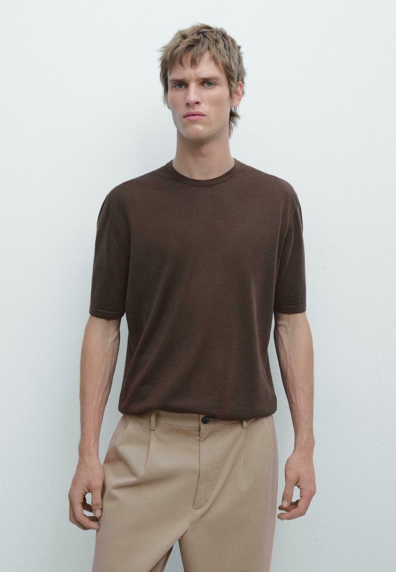 Jeune homme aux cheveux blonds courts, portant une chemise marron à manches courtes et un pantalon beige, debout devant un fond clair uni.