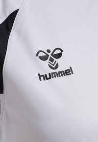 Gros plan sur un tissu de vêtements de sport blanc et noir présentant le logo noir de Hummel avec une abeille stylisée au-dessus du nom de la marque.